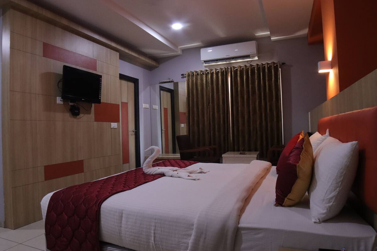 A hotel room with a bed, a flat-screen TV mounted on the wall, an air conditioning unit, and a curtain.
