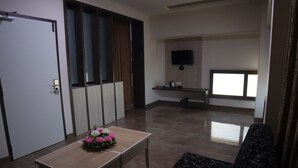 A modern living room with a sofa, a coffee table with flowers, a TV mounted on the wall, and a window with blinds. - Hotel PL A Residency Tiruchirappalli (Tiruchirappalli)