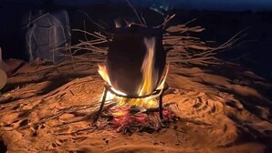 A campfire with a metal pot on a tripod stand, surrounded by logs and branches on a sandy ground. - Douz Trek (Douz Sud)