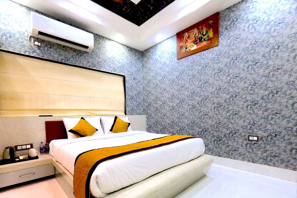 A modern hotel room with a large bed, a nightstand, a bedside table, an air conditioning unit, and a wall-mounted artwork.