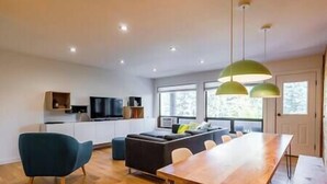 A modern living room with a wooden dining table, a sofa, and a TV. - Griz Inn Chic Condo - Mountain Views - Luxury Stay (Fernie)