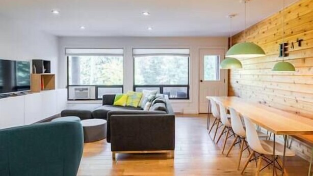 A modern living room with a sofa, a dining table with chairs, and a kitchen area with a wooden wall and green pendant lights. - Griz Inn Chic Condo - Mountain Views - Luxury Stay (Fernie)