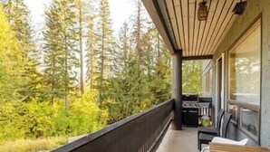 A balcony with a view of dense forest, a grill, and a chair. - Griz Inn Chic Condo - Mountain Views - Luxury Stay (Fernie)