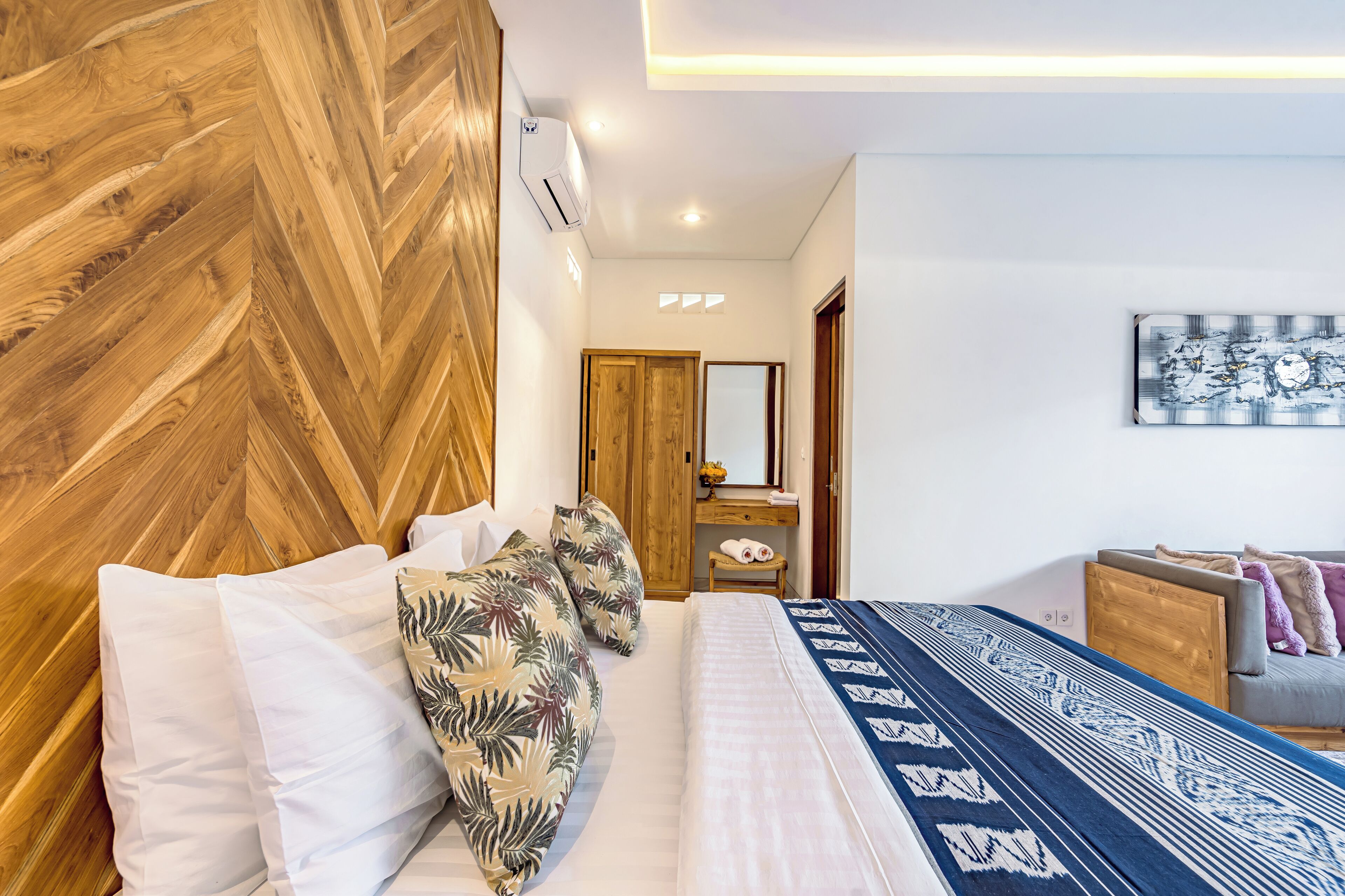 A hotel room with a wooden headboard, a bed with white and patterned bedding, a small wooden nightstand, a mirror, and a gray sofa with cushions.