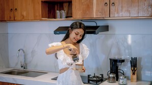 A kitchen with wooden cabinets, a sink, a coffee maker, and a person pouring coffee. - Sapta D'Villa Residence by Purely (Sukawati)