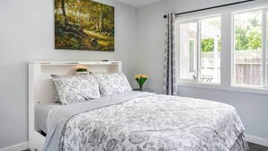 A bedroom with a bed, a painting, a window with curtains, and a small table with flowers. - Adorable House with EV&Parking-Family Friendly (Culver City)