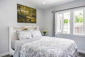 Un dormitorio con cama, un cuadro, una ventana con cortinas y una pequeña mesa con flores.