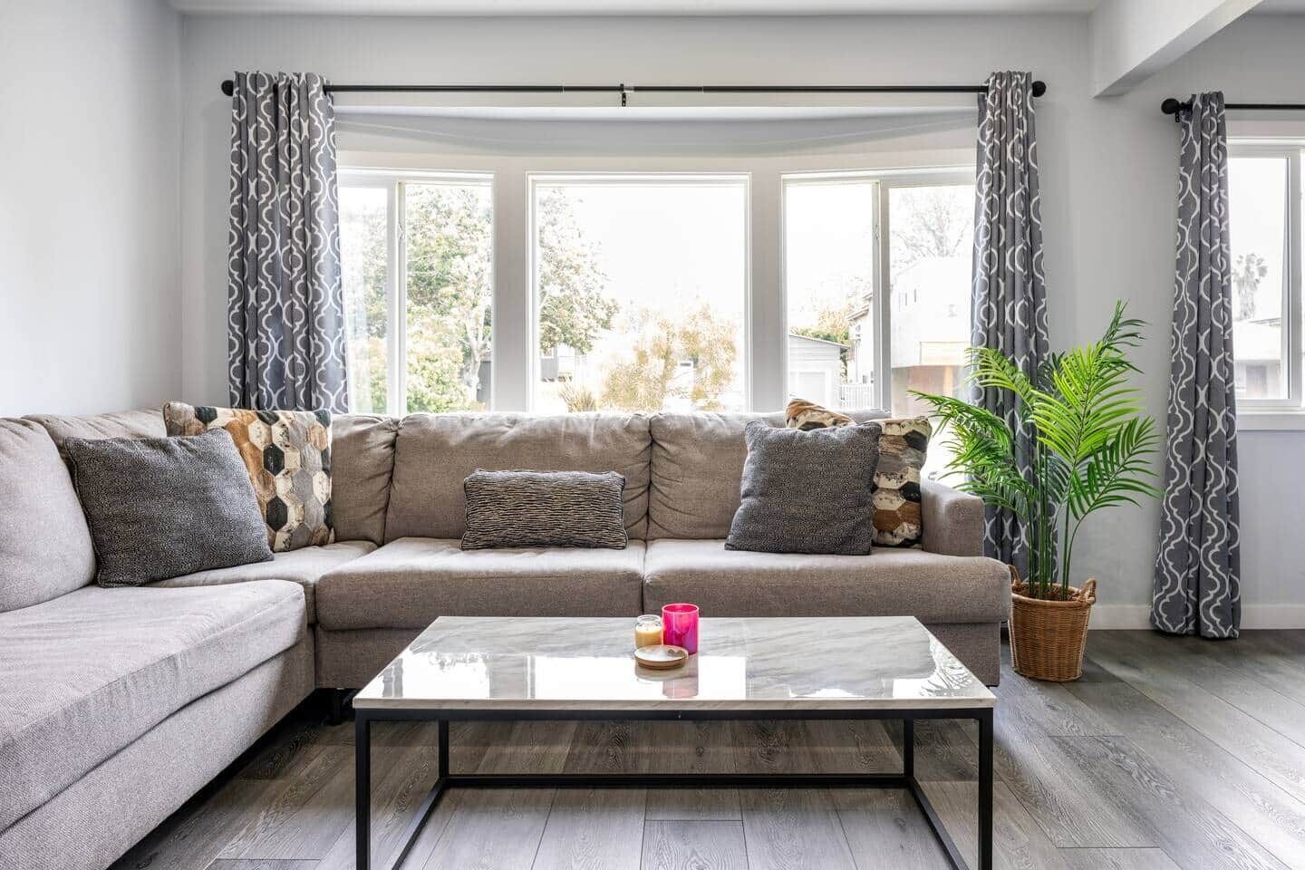 En stue med en grå sofa, mønstrede puder, et glas sofabord og en potteplante.