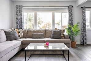 Una sala de estar con un sofá gris, cojines estampados, una mesa de centro de vidrio y una planta en maceta.