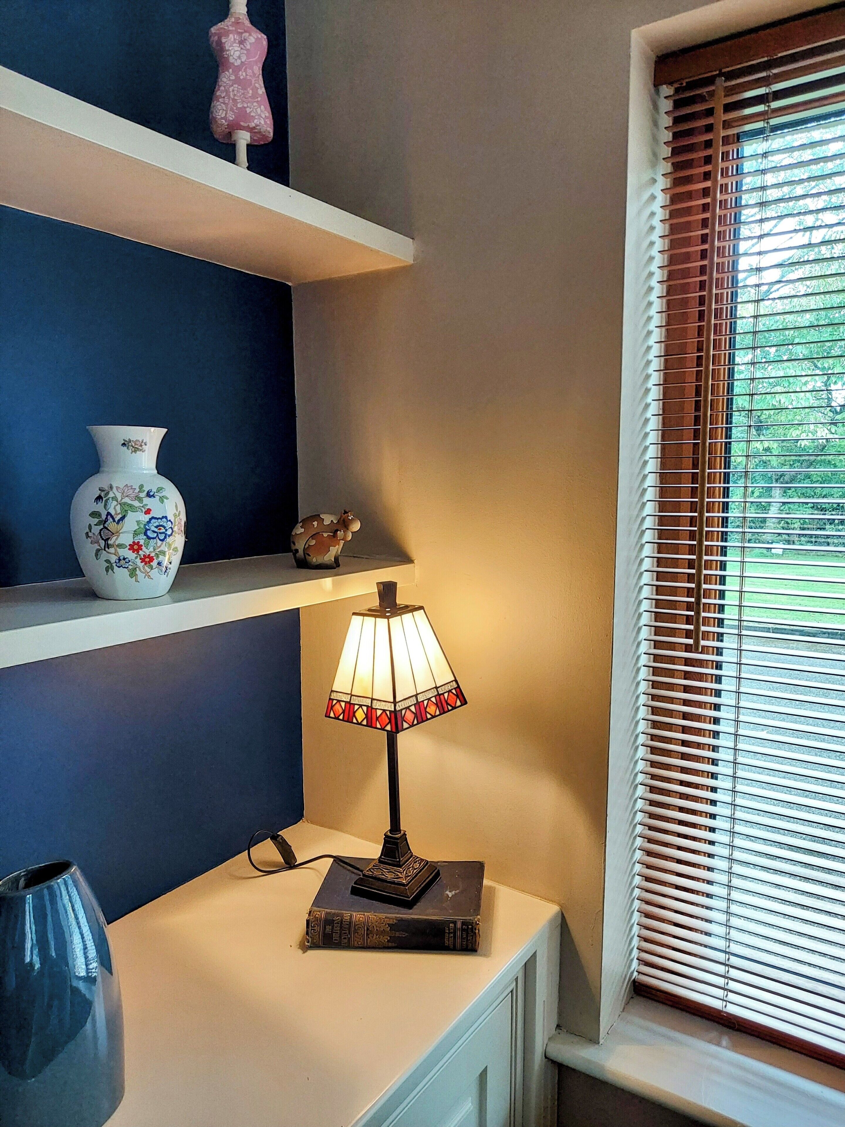 A room with a white radiator, a lamp on a small table, a vase, and a window with blinds.
