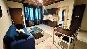 A modern living room with a blue sofa, a coffee table, and a dining area with a table set for two. - OMA Residence (Pavia)