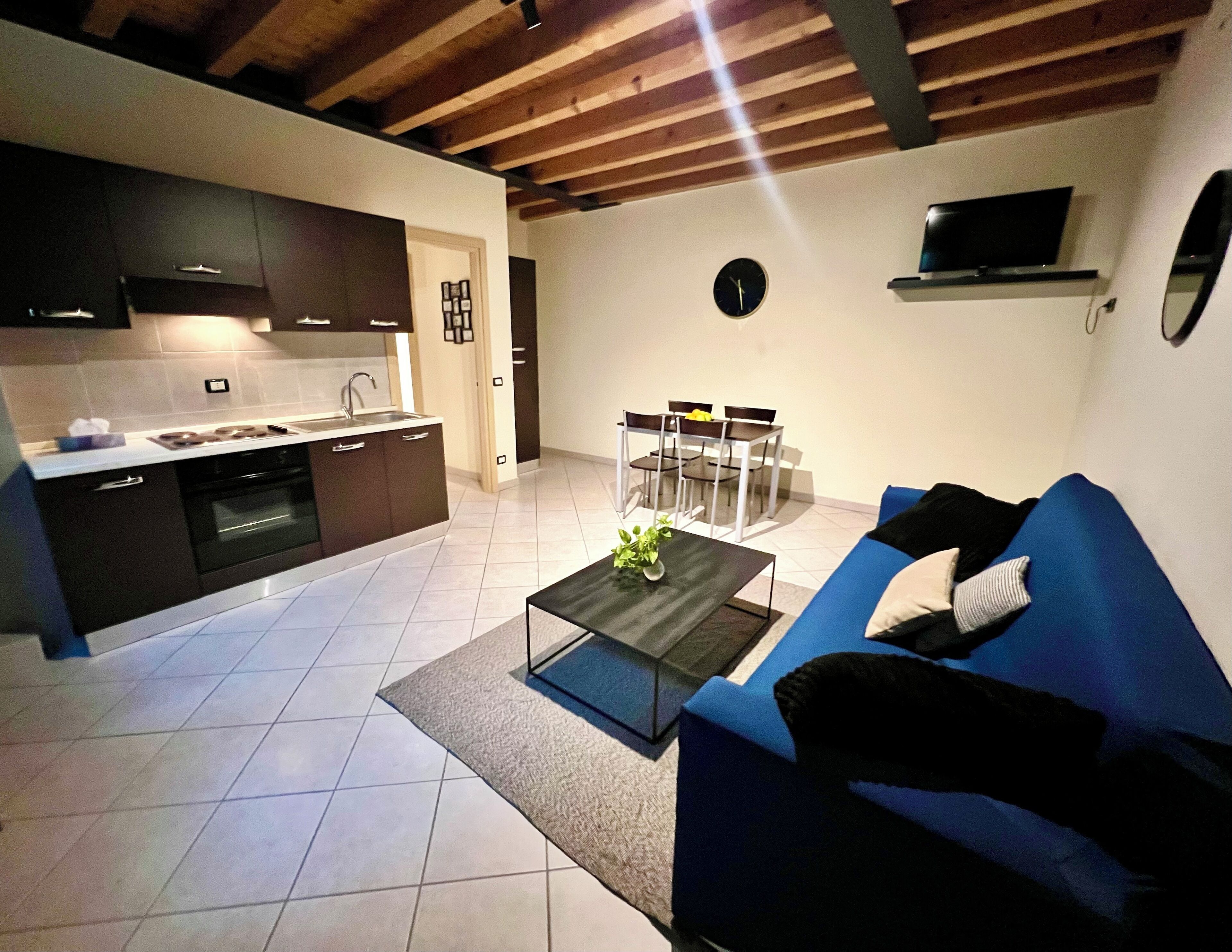 A modern kitchen with dark cabinets, a dining area with a table and chairs, and a living room with a blue sofa.