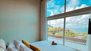 Un dormitorio con un ventanal grande, una cama con almohadas blancas y amarillas, y vistas a una zona costera con edificios y una vía de tren.