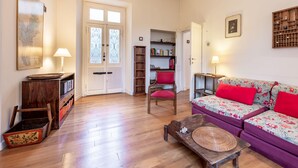 Ein Wohnzimmer mit Holzboden, einem violetten Sofa mit roten Kissen, einem Holzstuhl, einem Bücherregal und einer Tür mit Milchglasfenstern.