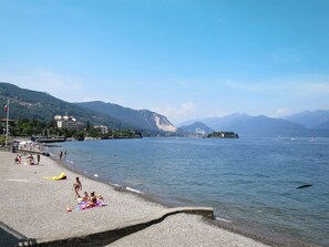 A pebbled beach with people sunbathing and playing, a clear blue sky, and mountains in the background. - Delle Ortensie by Interhome (Stresa)