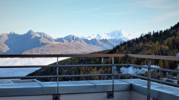 Un balcón con barandilla de metal que da a un paisaje montañoso con picos nevados y un valle boscoso.