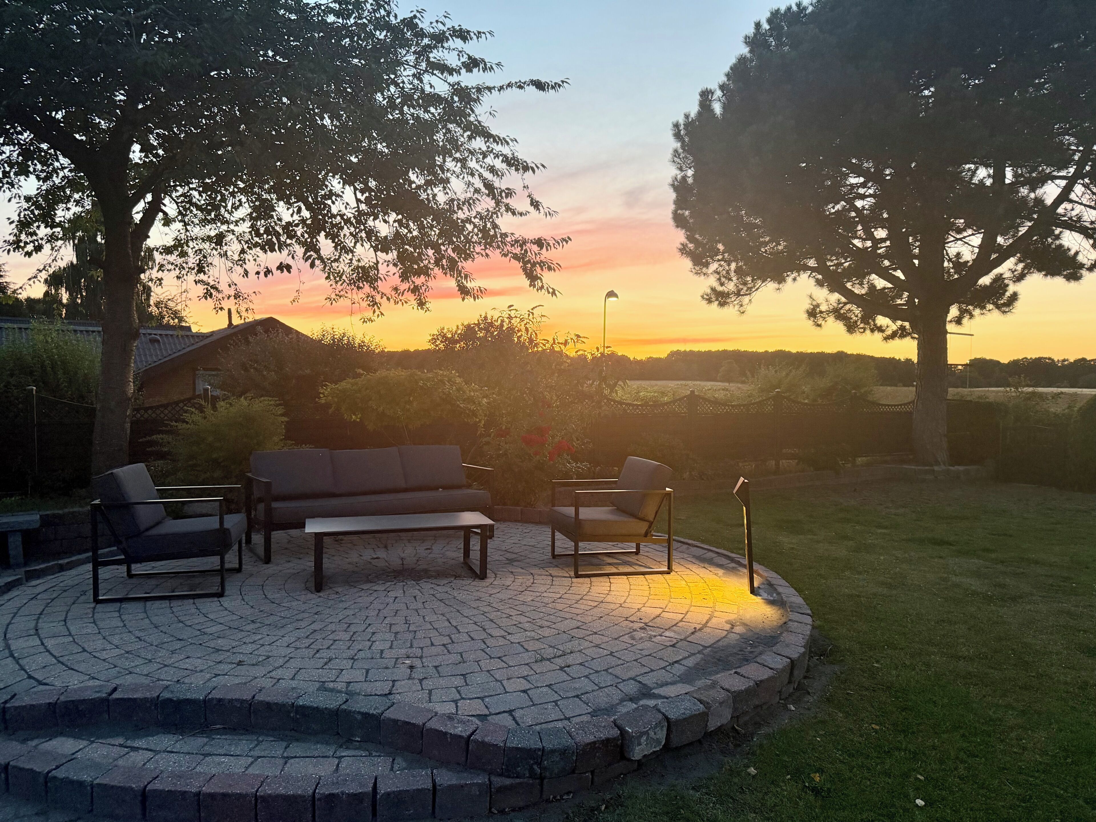 A circular outdoor seating area with a brick floor, surrounded by trees and a sunset view.