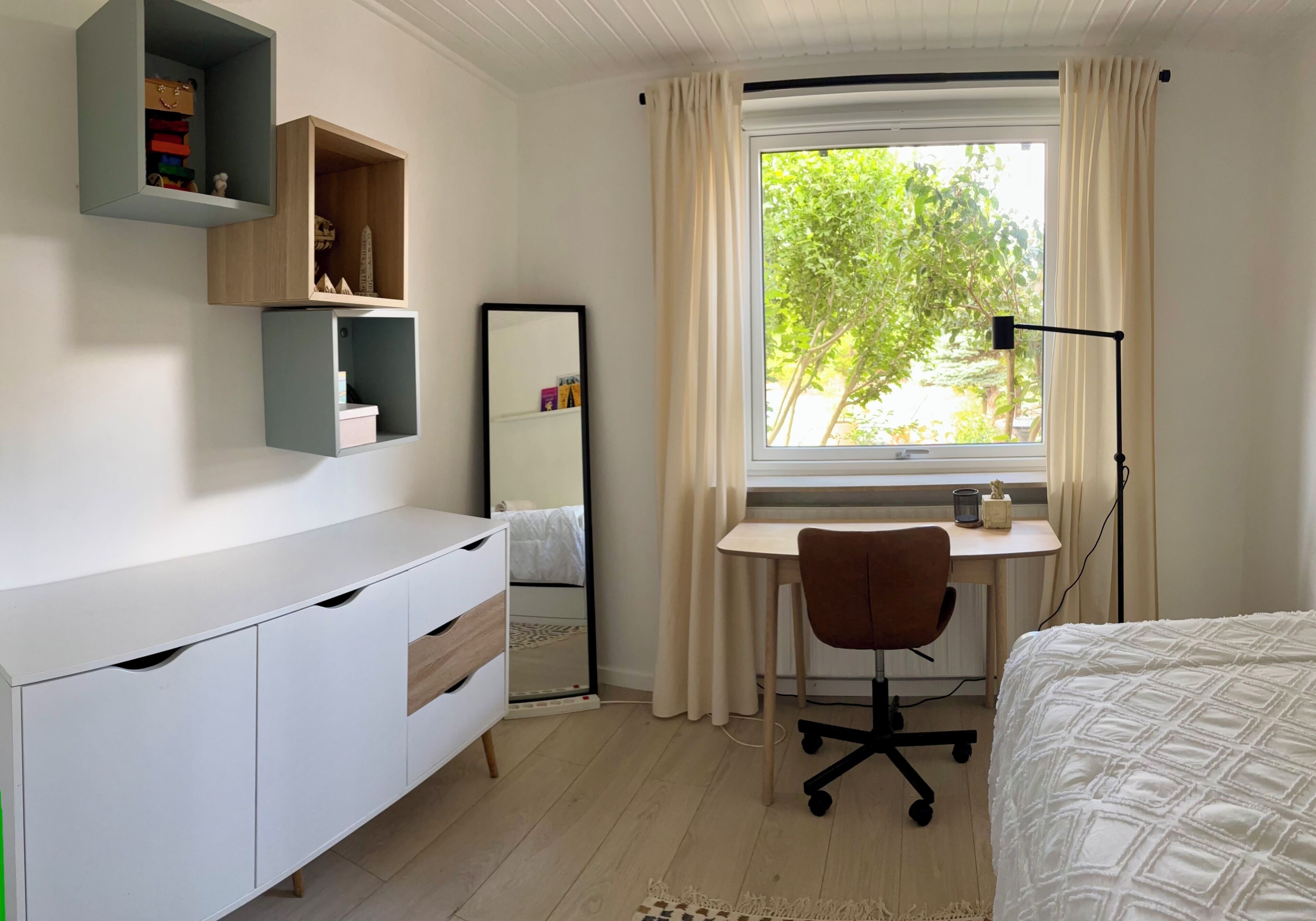 A bedroom with a bed, a desk, a chair, a wardrobe, a mirror, and a window with curtains.