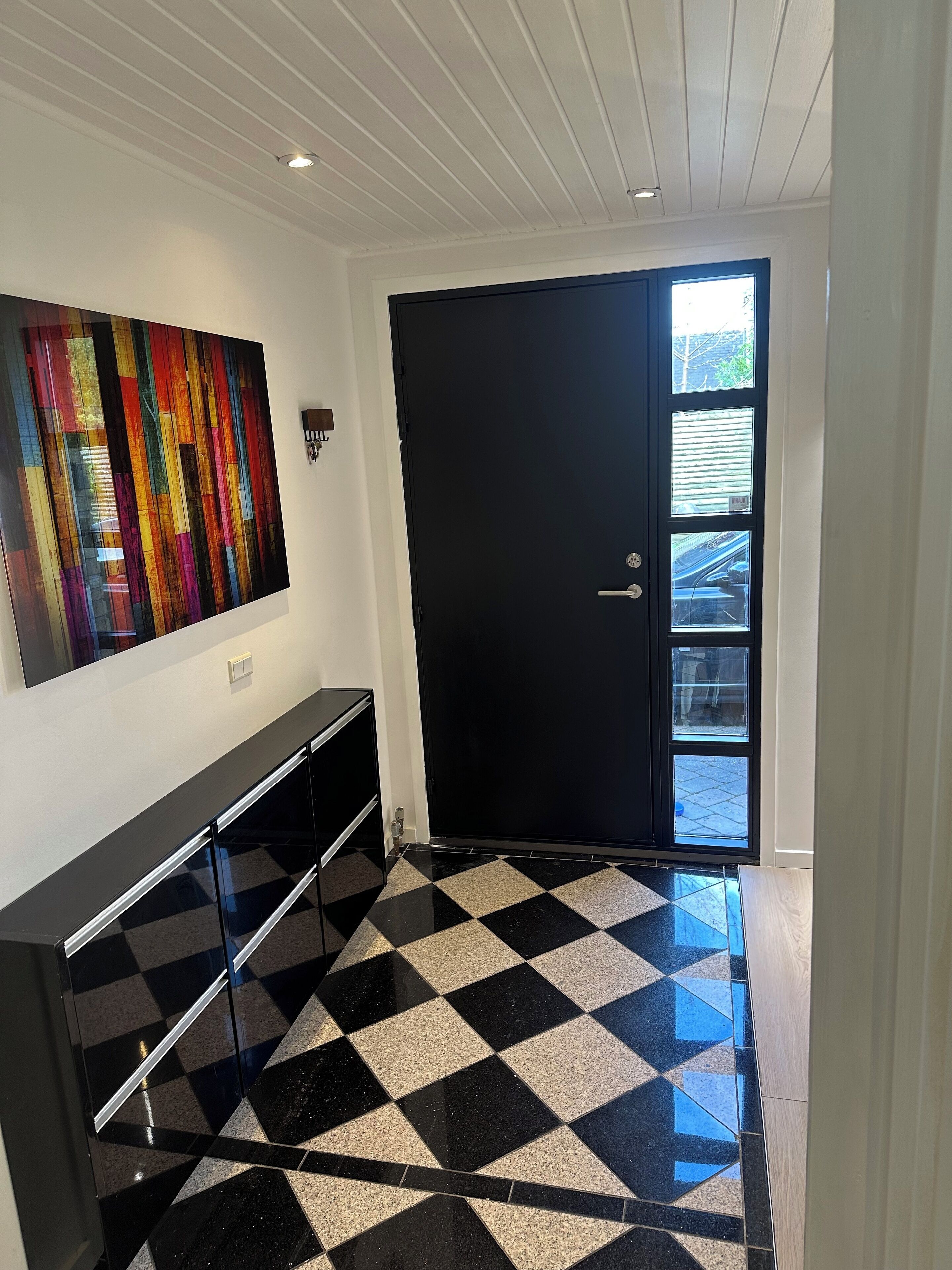 Un intérieur moderne avec un sol à carreaux noirs et blancs, une porte noire dotée d’une fenêtre et un tableau abstrait aux couleurs vives accroché au mur.