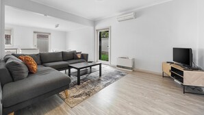 A modern living room with a grey sectional sofa, a coffee table, a TV on a wooden stand, and a large window with a view of greenery.