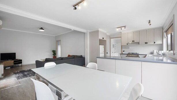 A modern kitchen with white cabinets, a dining area with a white table and chairs, and a living area with a sofa and a television.