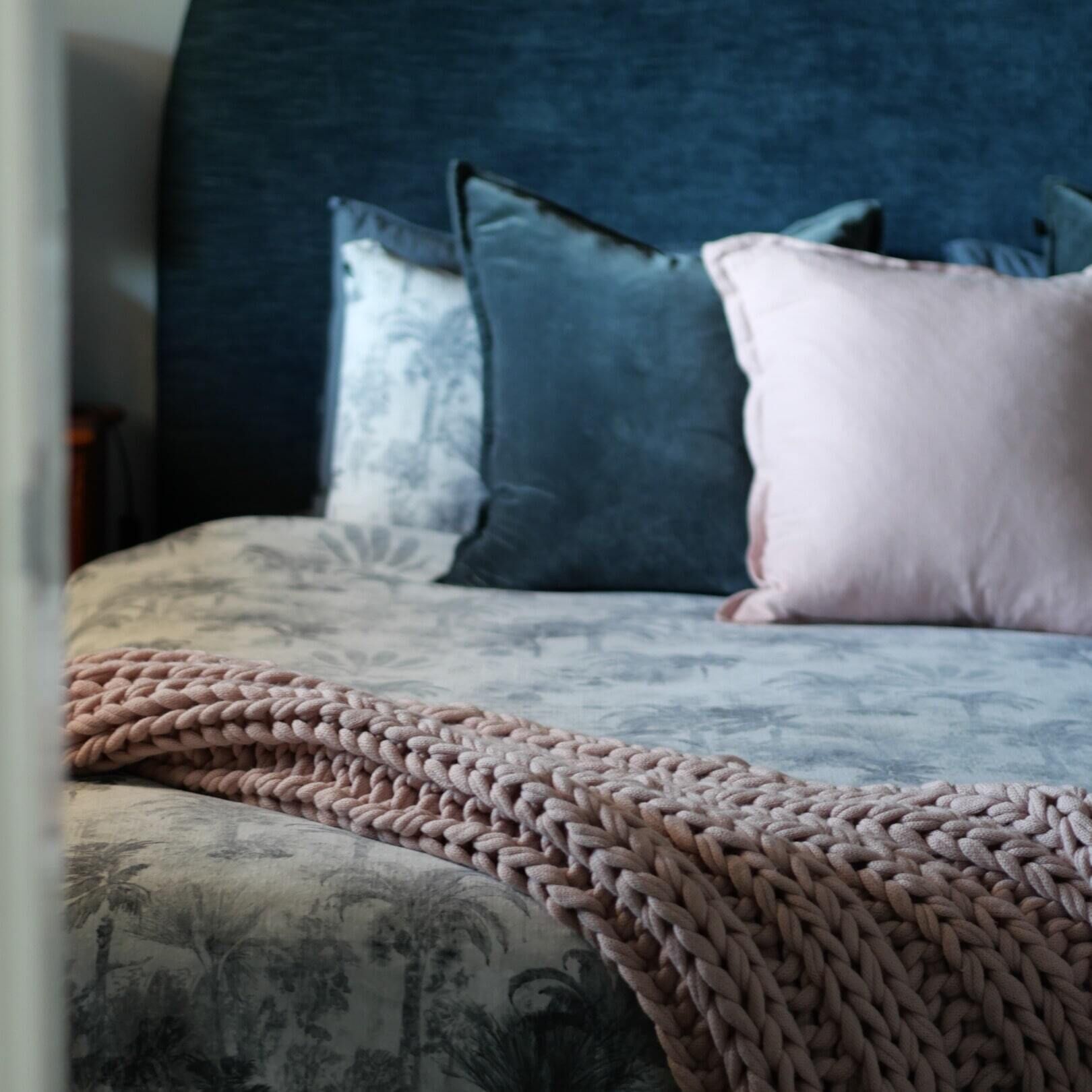 A bed with a patterned duvet cover, a knitted throw blanket, and multiple pillows in shades of blue and pink.