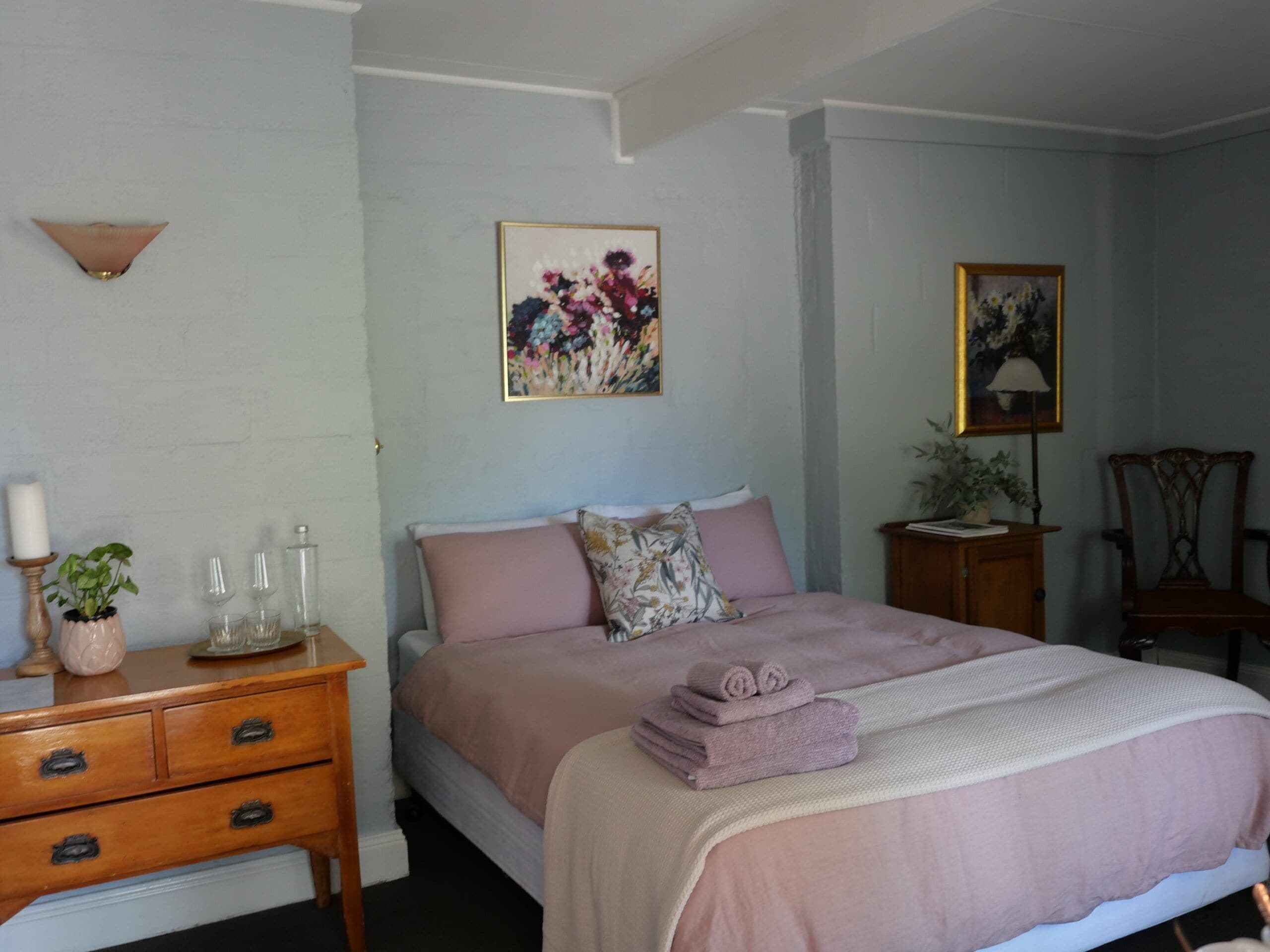 Un dormitorio con cama, cómoda, mesita de noche, lámpara de pared y un cuadro con motivos florales.