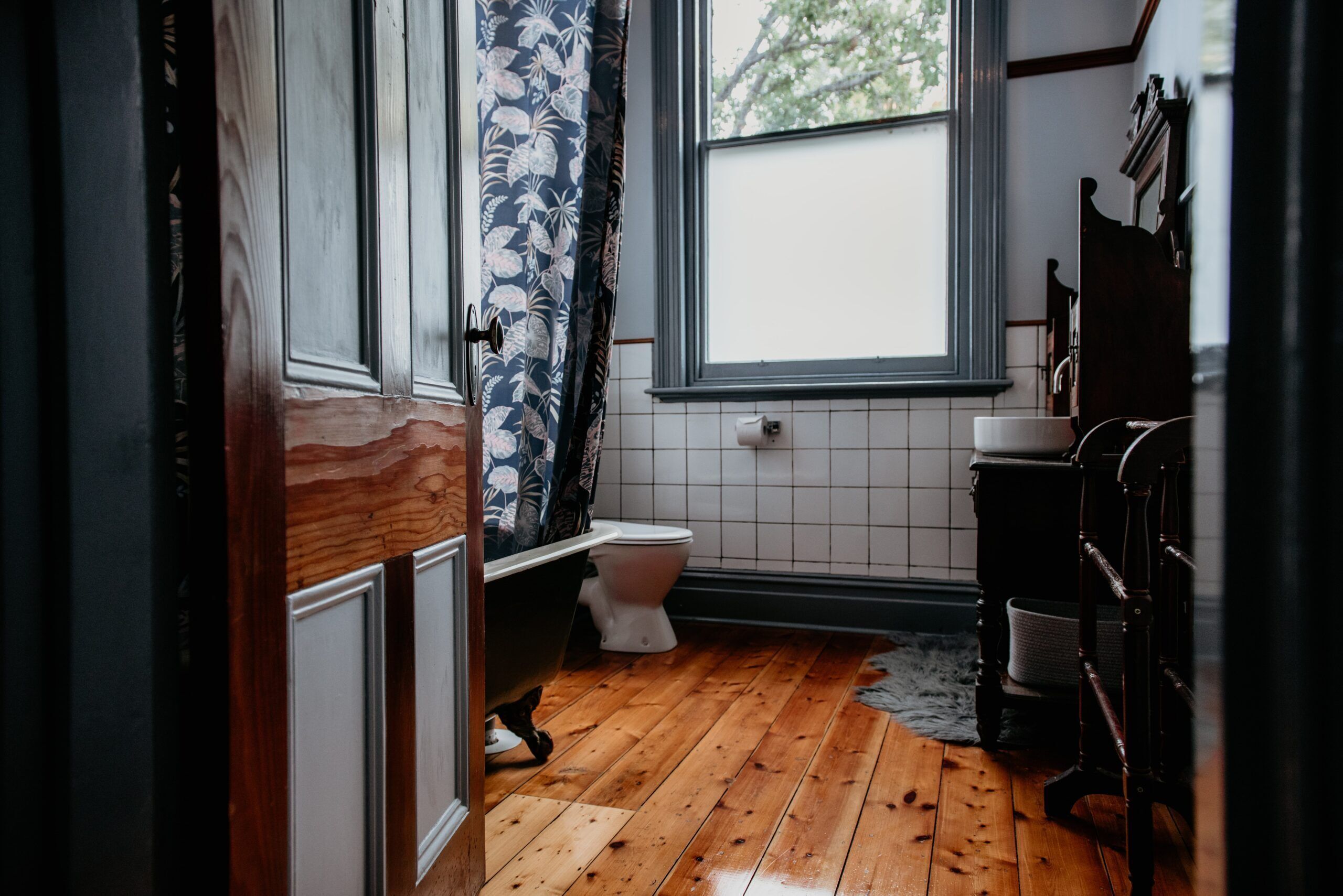 Une salle de bain avec un plancher en bois, des carreaux blancs, des toilettes, un lavabo et une fenêtre donnant sur des arbres.