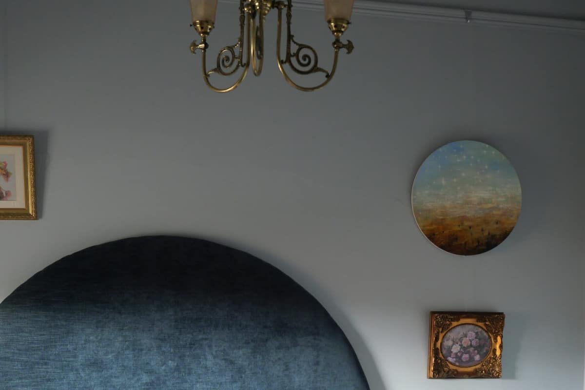 Quarto com uma grande cabeceira azul escura, um relógio de parede redondo, um quadro emoldurado e uma mesa de cabeceira com abajur.