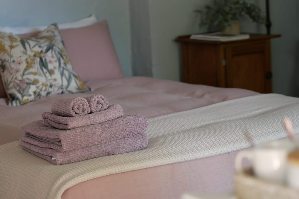 Cama bem arrumada com toalhas cor-de-rosa, um cobertor branco e uma mesinha de cabeceira de madeira com abajur e um livro.