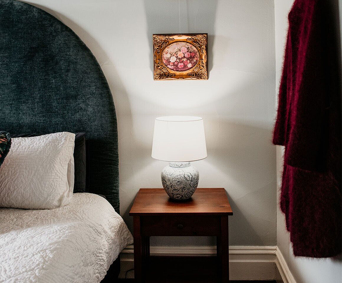 Un dormitorio con cama, una mesita de noche con lámpara, un cuadro enmarcado en la pared y una toalla roja colgada.