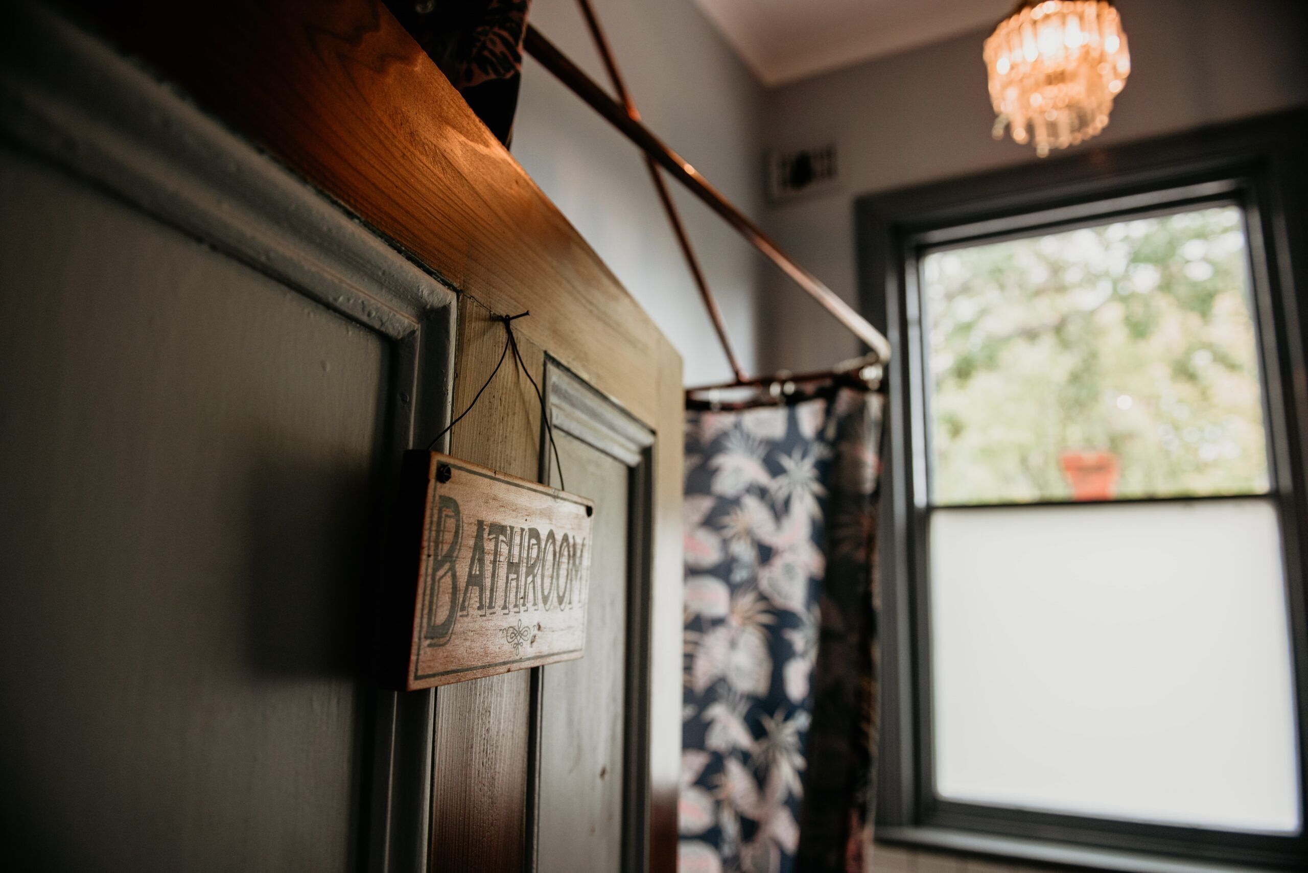 A wooden sign labeled "BATHROOM" on a door, a chandelier, and a window with a view of greenery.