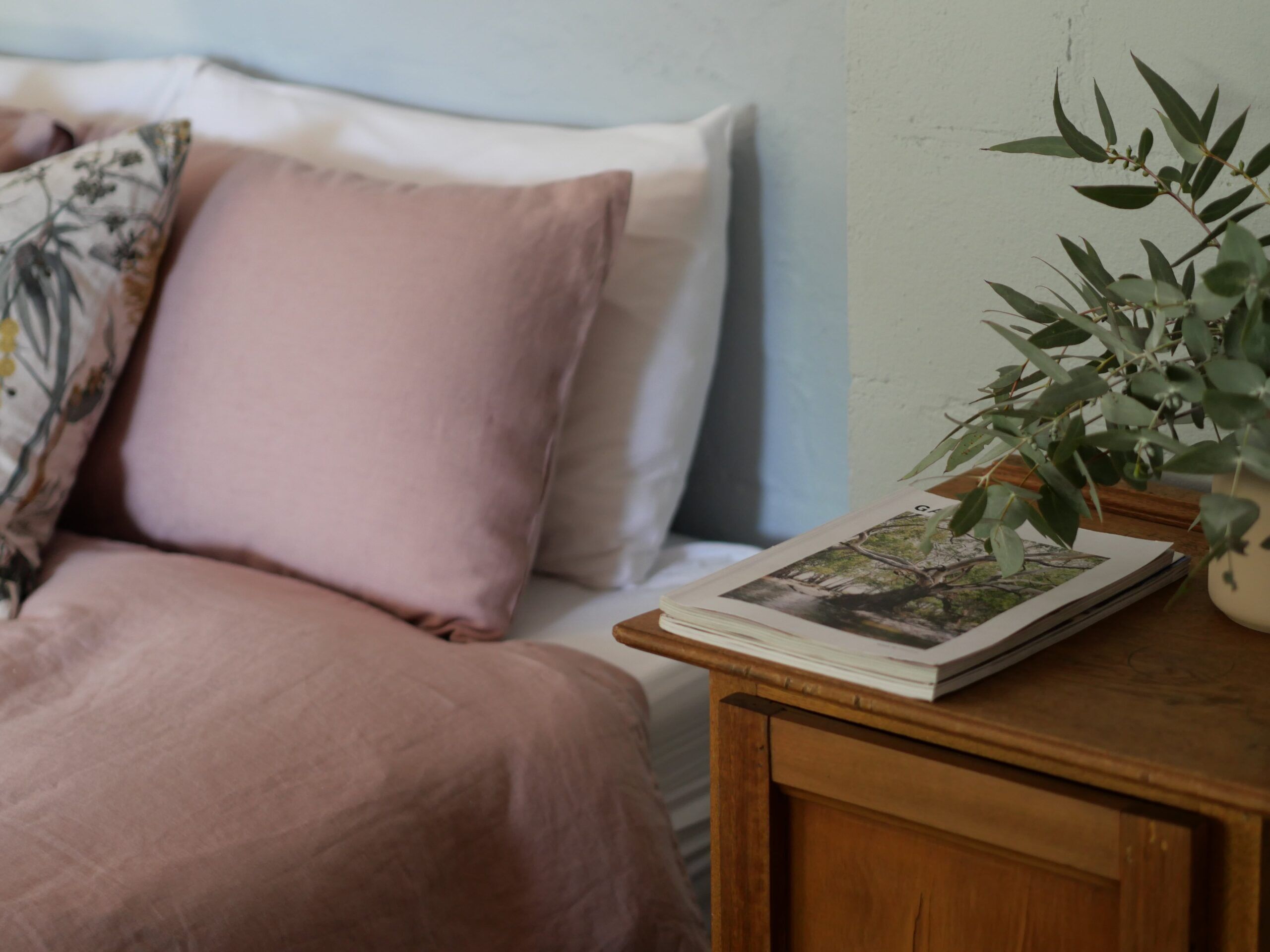 A neatly made bed with a pink blanket and pillows, a wooden bedside table with a book and a potted plant.