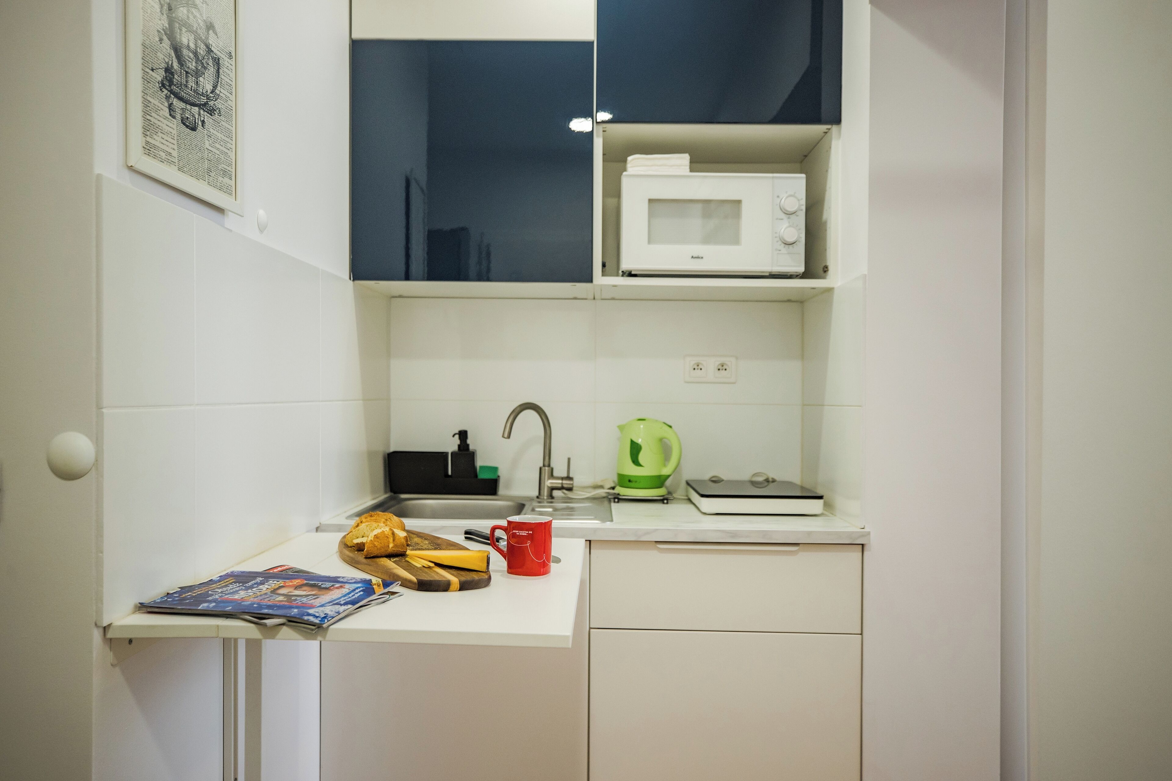 A compact kitchenette with a microwave, kettle, sink, and a small countertop with a magazine and a red mug.