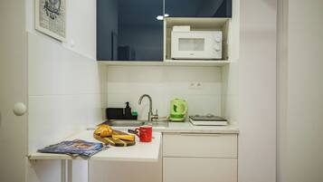 A compact kitchenette with a microwave, kettle, sink, and a small countertop with a magazine and a red mug.