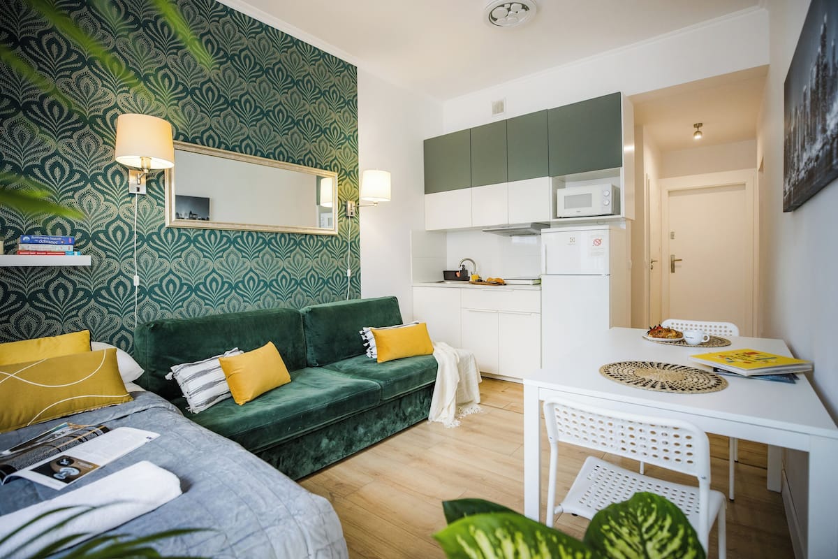Um espaço de estar compacto com uma parede estampada em verde, um sofá verde com almofadas amarelas, uma mesa branca e uma kitchenette com micro-ondas.