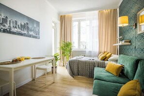 A modern hotel room with a bed, a green sofa, a dining table with chairs, and a cityscape painting on the wall. - MGR Studio Bitwy Warszawskiej Warszawa (Warsaw)
