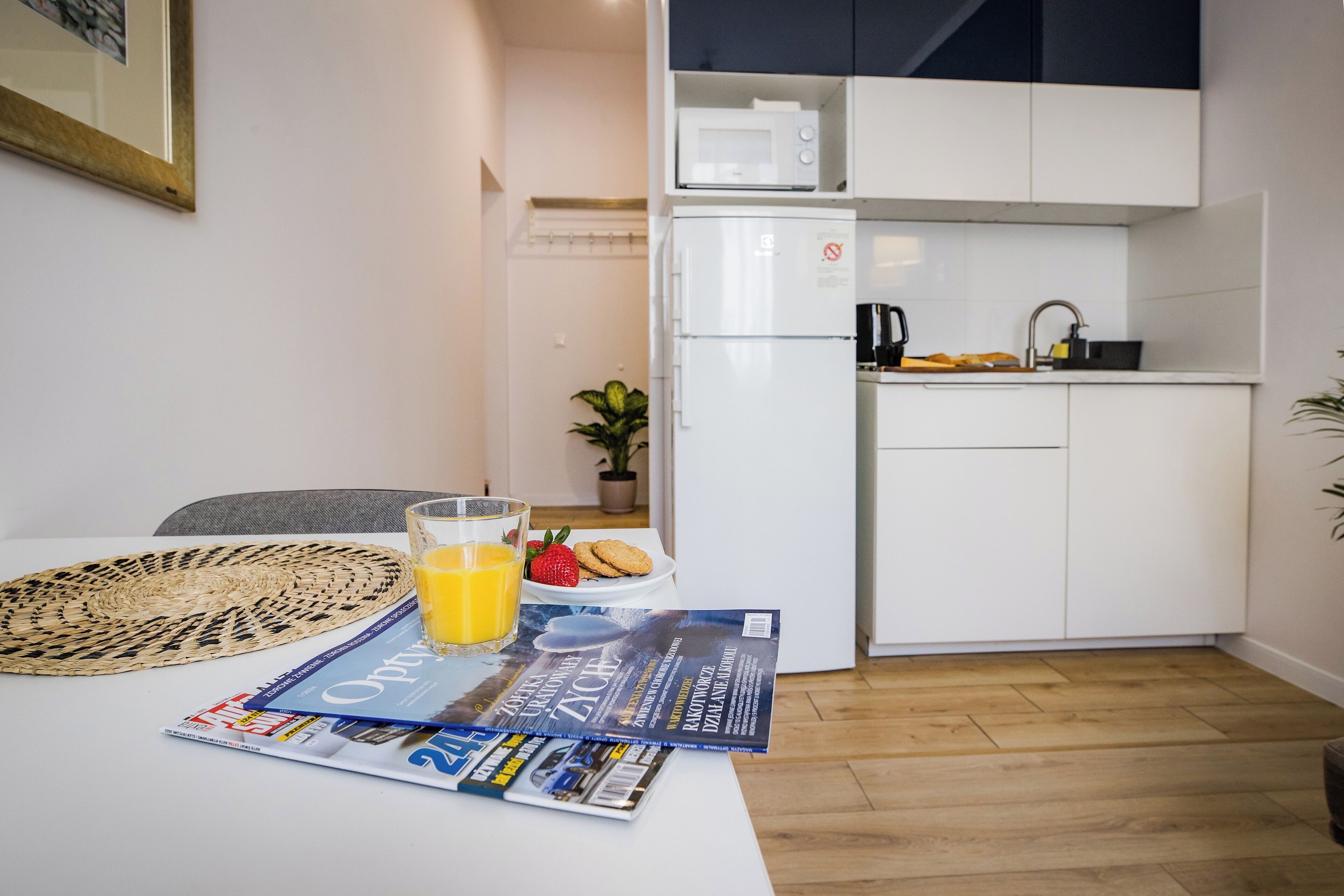 Une cuisine moderne avec des meubles blancs, un four à micro-ondes et un réfrigérateur. On y trouve une table à manger avec un magazine, un verre de jus d’orange et une assiette contenant des biscuits et une fraise.