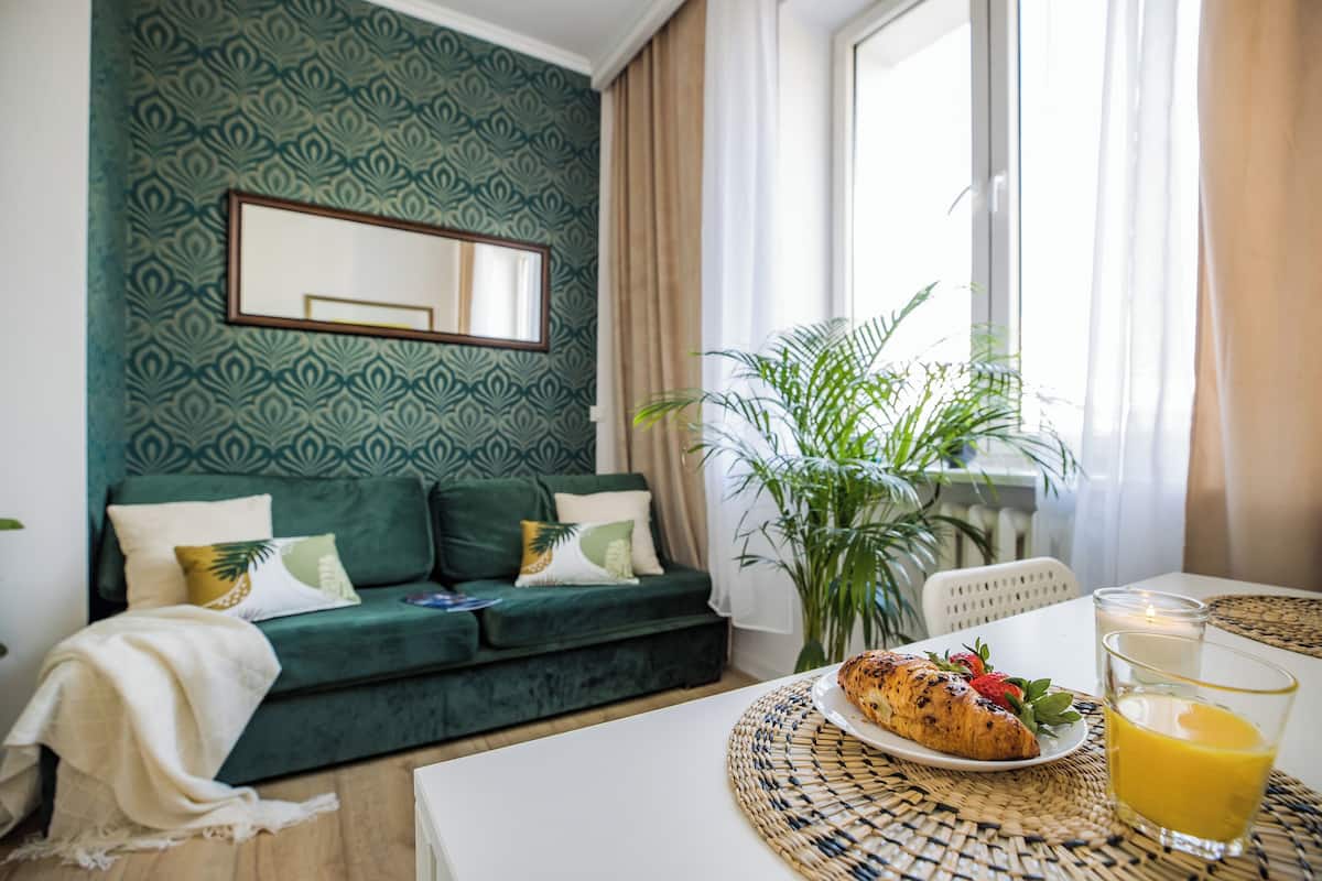 Uma sala de estar acolhedora com parede estampada em verde, um sofá com almofadas, uma mesa de jantar com um prato de comida e um copo de suco, e uma janela grande com cortinas.