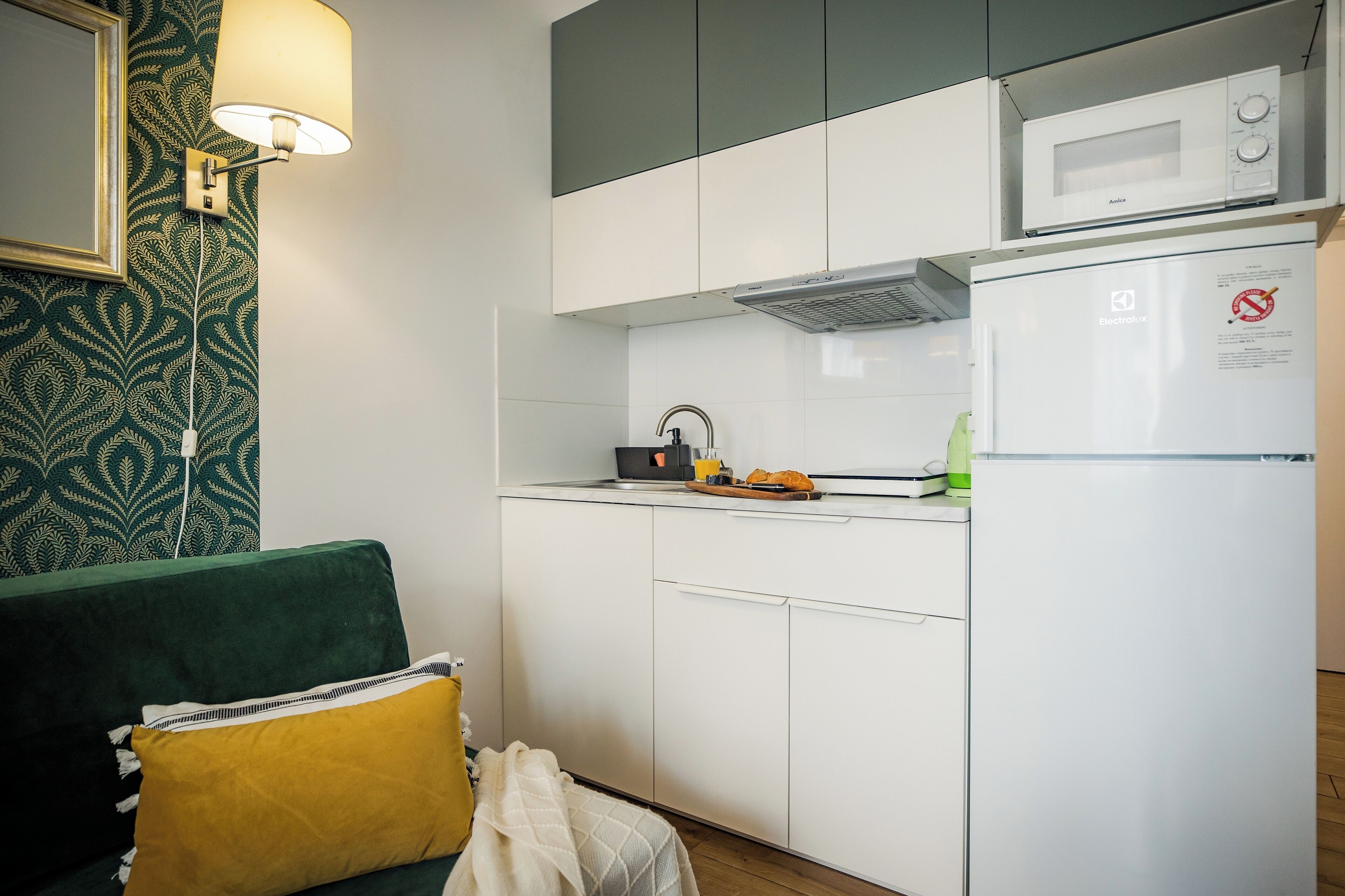 Une cuisine compacte équipée d’un four à micro-ondes, d’une bouilloire et d’un grille-pain. On y trouve un canapé vert avec un coussin jaune et une couverture blanche.
