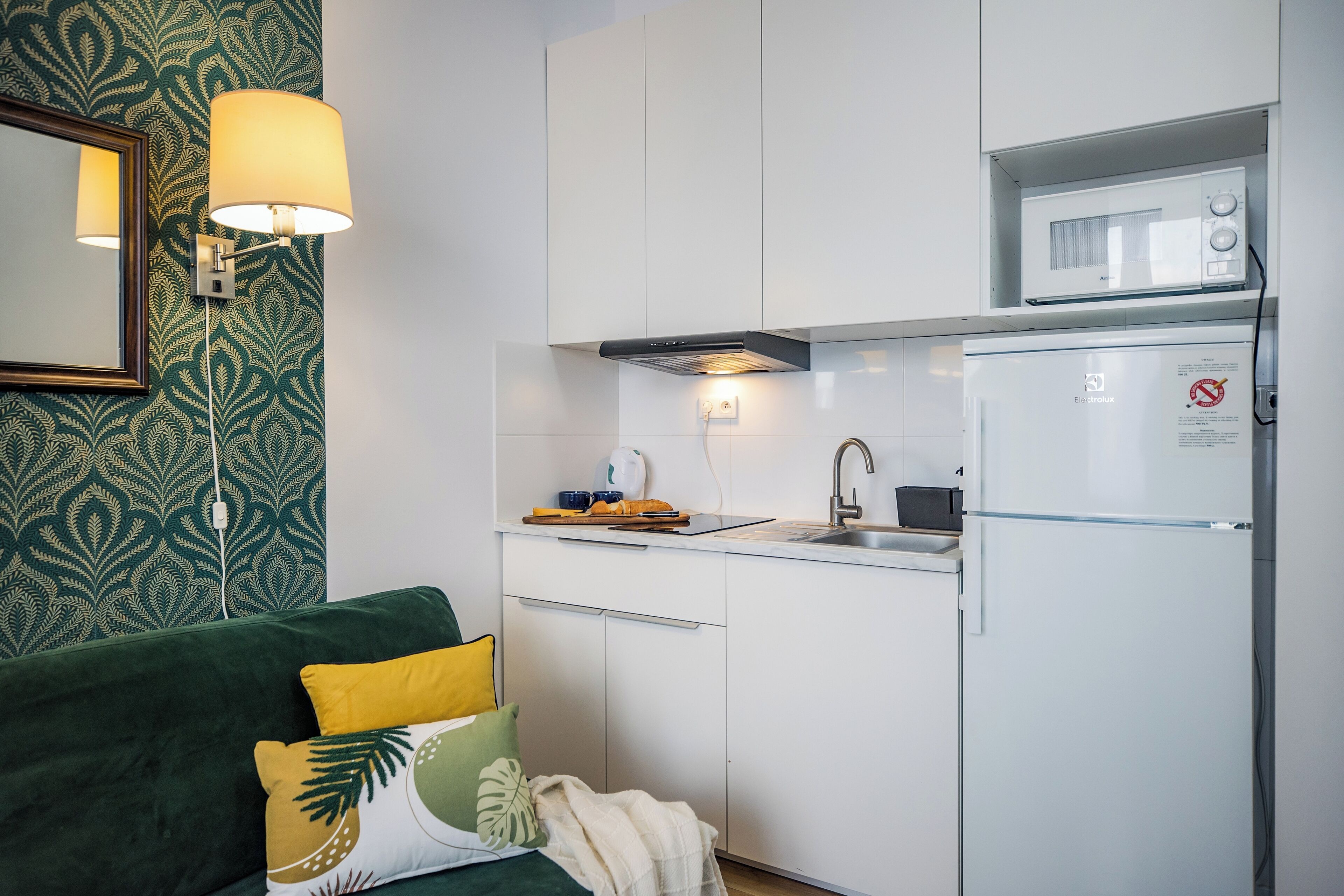 A compact kitchen with a microwave, a sink, and a refrigerator. There is a green couch with decorative pillows and a wall-mounted lamp.