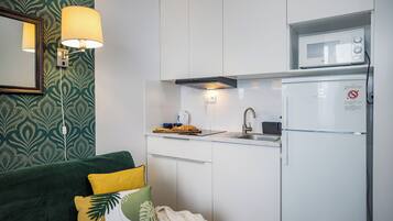 A compact kitchen with a microwave, a sink, and a refrigerator. There is a green couch with decorative pillows and a wall-mounted lamp.