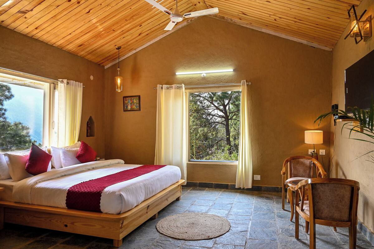 Un dormitorio con techo de madera, una cama con ropa de cama blanca y almohadas rojas, suelo de piedra, alfombra redonda, una silla de madera, una mesa, una ventana con cortinas y vistas a árboles fuera.
