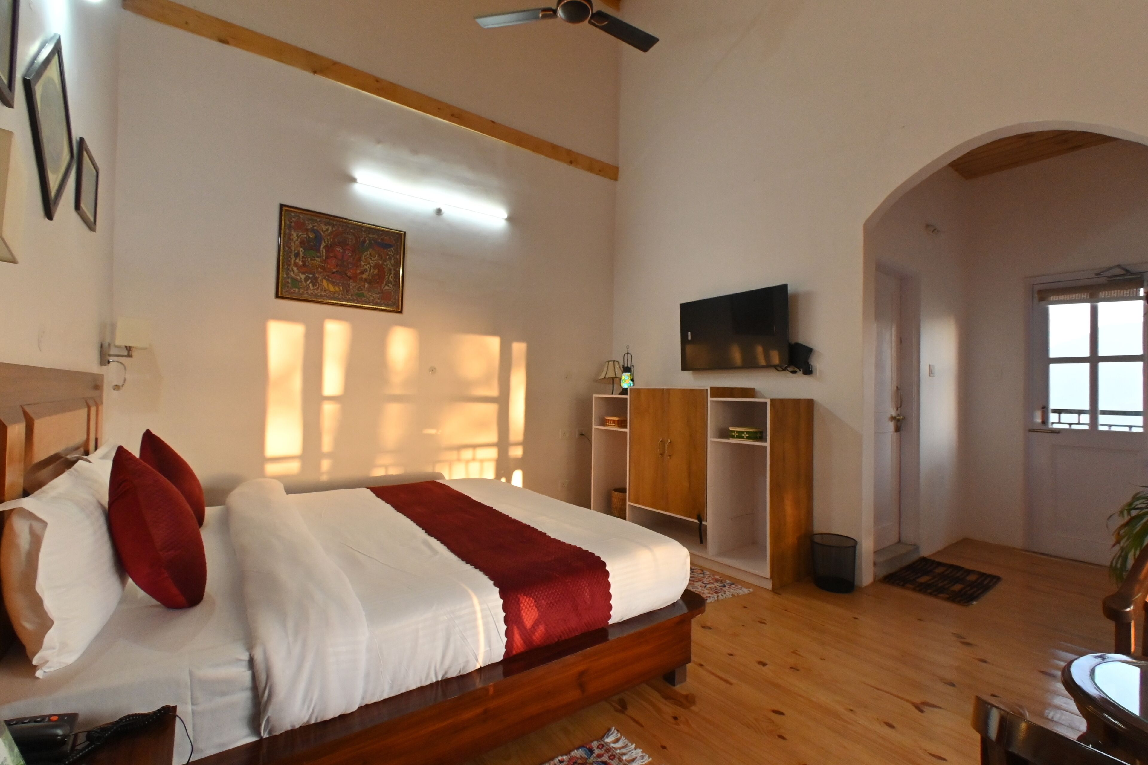 A bedroom with a bed, wooden floor, a ceiling fan, a TV on the wall, and a door leading to another room.