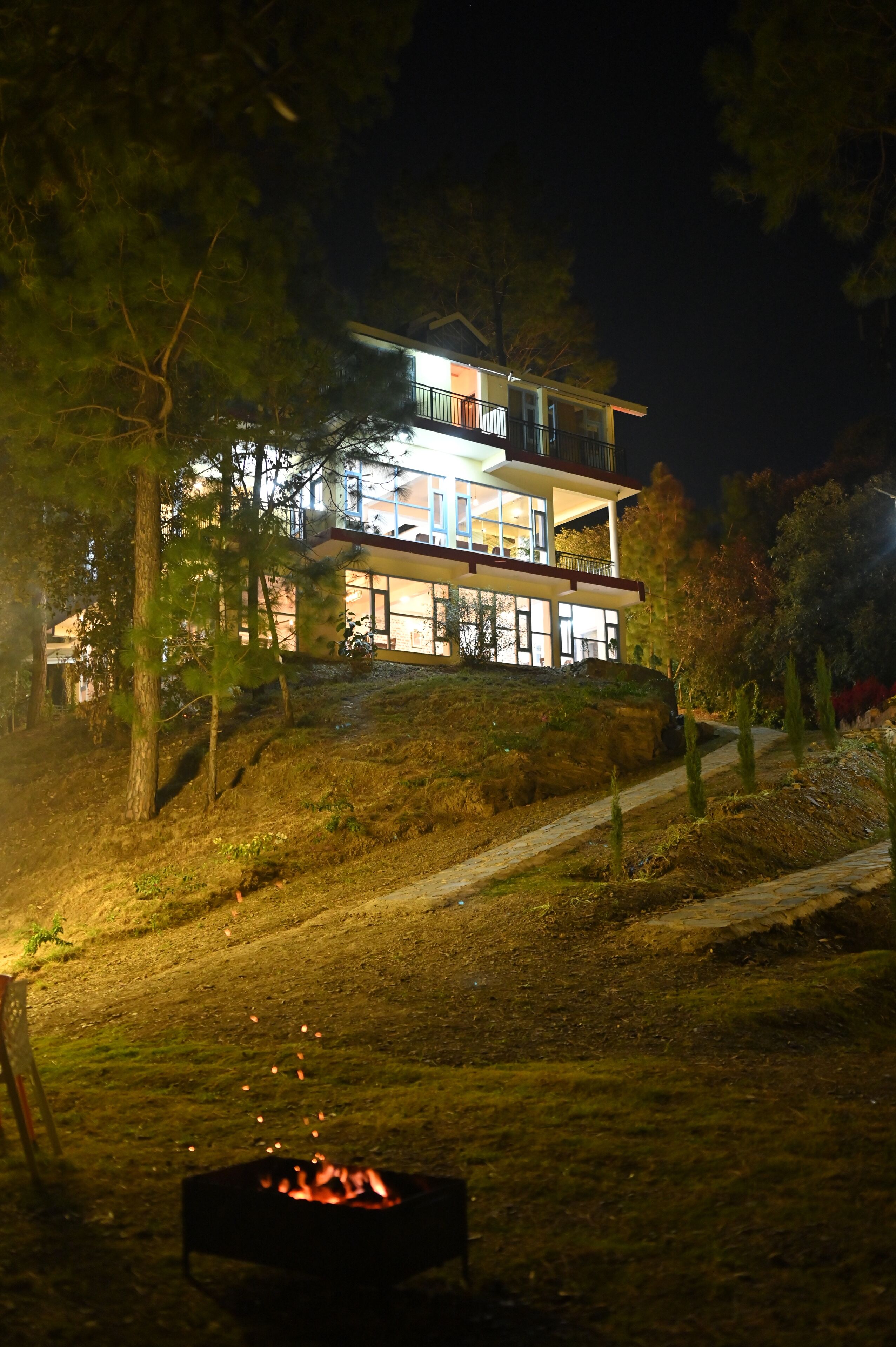 A multi-story house with a balcony, surrounded by trees and a fire pit in the foreground.