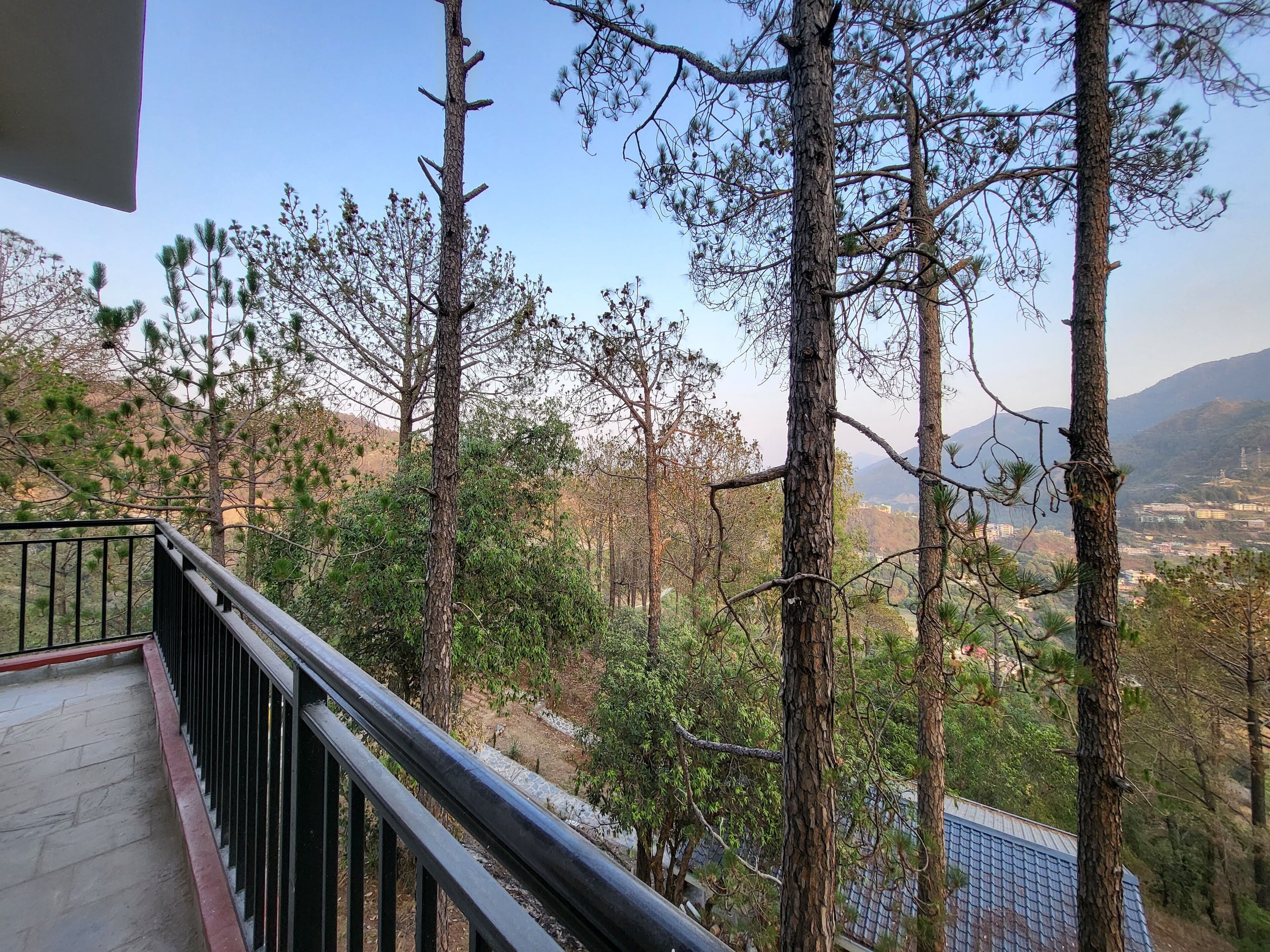 A balcony with a metal railing overlooking a mountainous landscape with trees and a distant view of buildings.