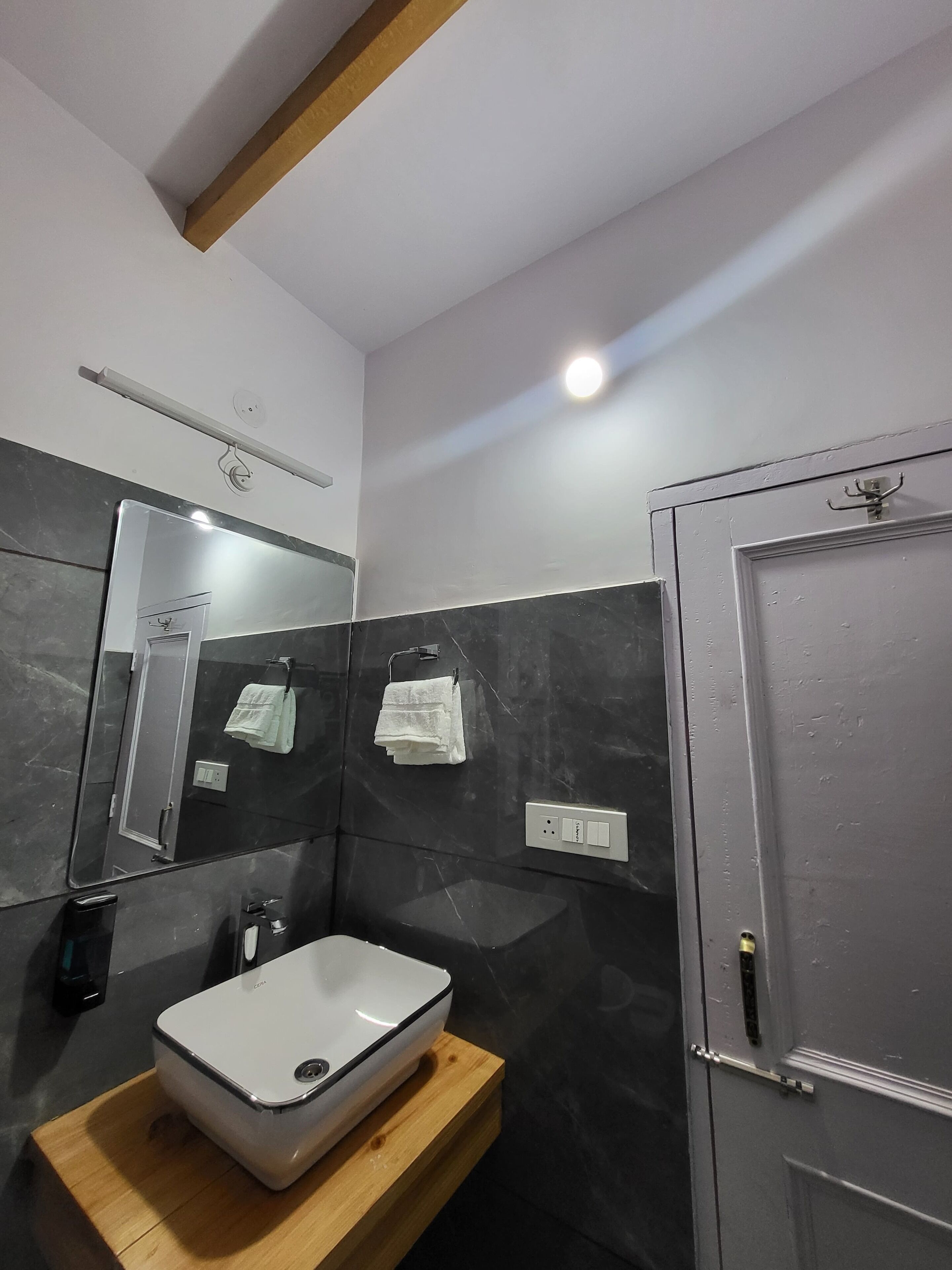 Une salle de bain moderne avec un lavabo blanc, des murs recouverts de carreaux foncés, un meuble-lavabo en bois, un grand miroir et une porte dotée d’une poignée.