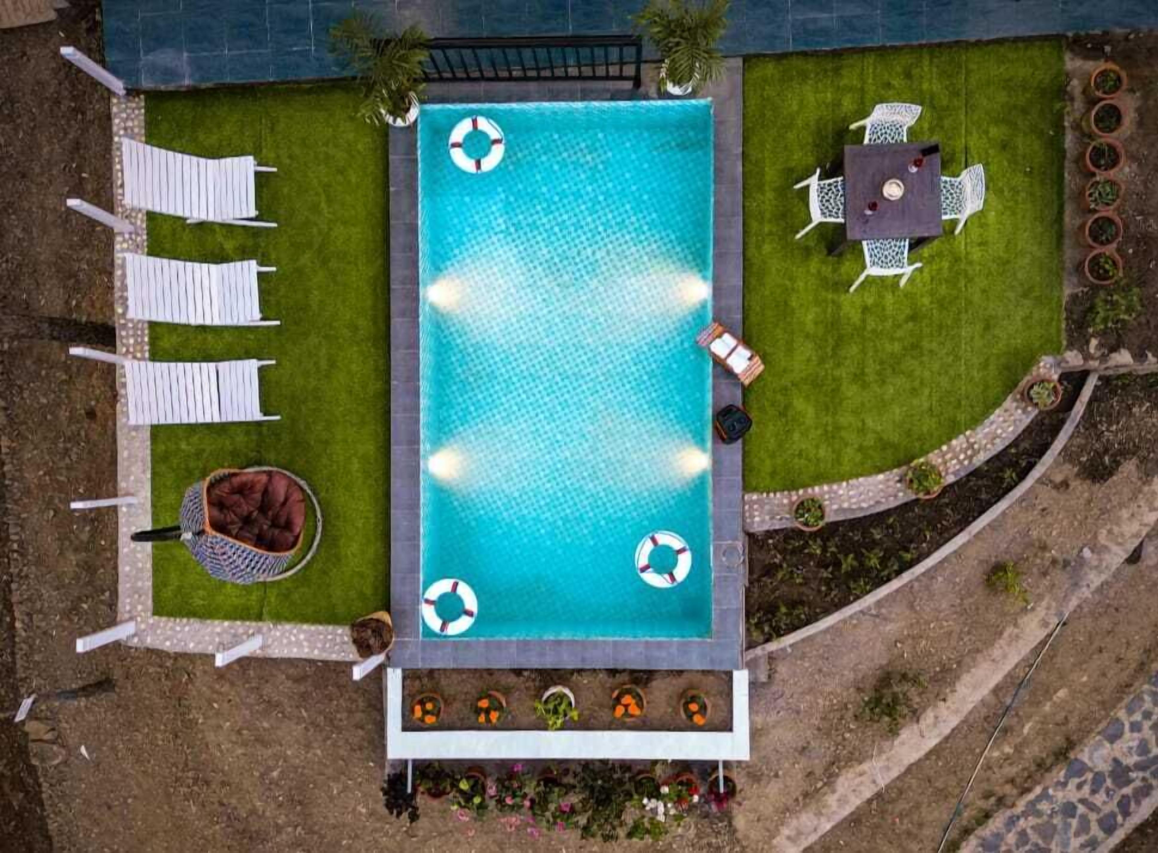 An aerial view of a rectangular swimming pool with a lifebuoy, surrounded by white lounge chairs, a table with chairs, and a small garden area with potted plants.