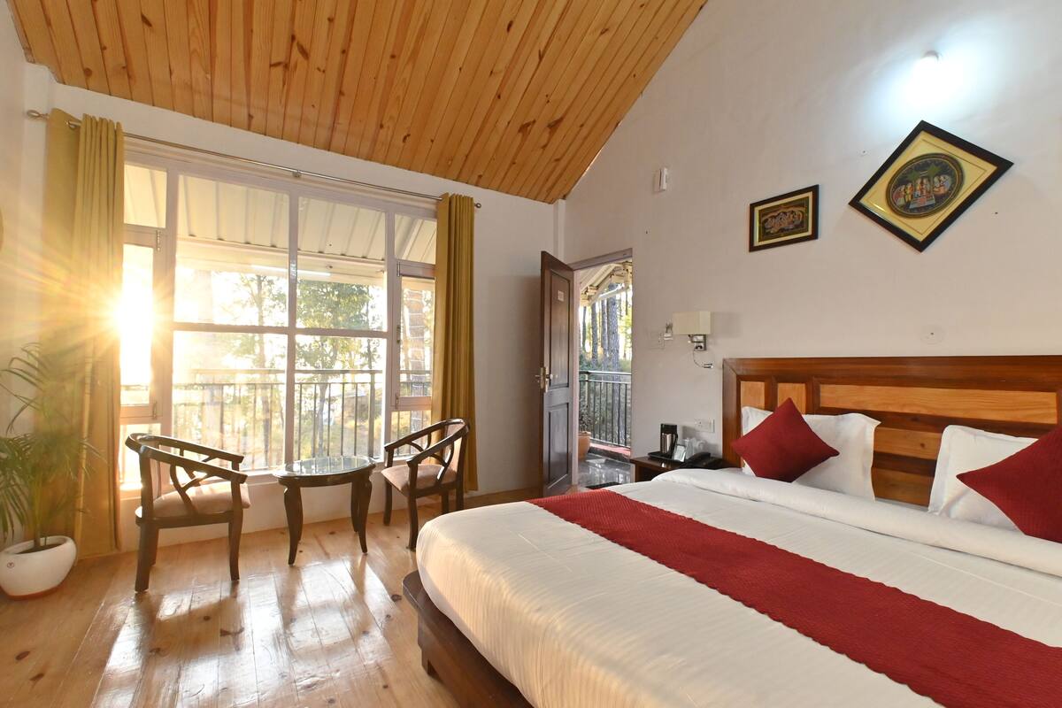 Habitación de hotel con techo de madera, una cama grande con ropa de cama roja y blanca, una mesita con sillas y una ventana con cortinas.