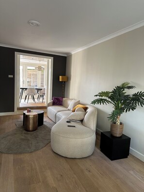 A modern living room with a sectional sofa, a round ottoman, a potted plant, and a dining area visible through a glass door.